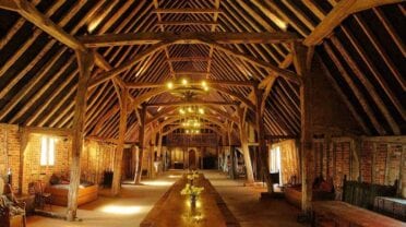 The Tudor Barn, Suffolk – Historic timber-frame barn interior with exposed oak beams and brick walls, lit by uplighting.