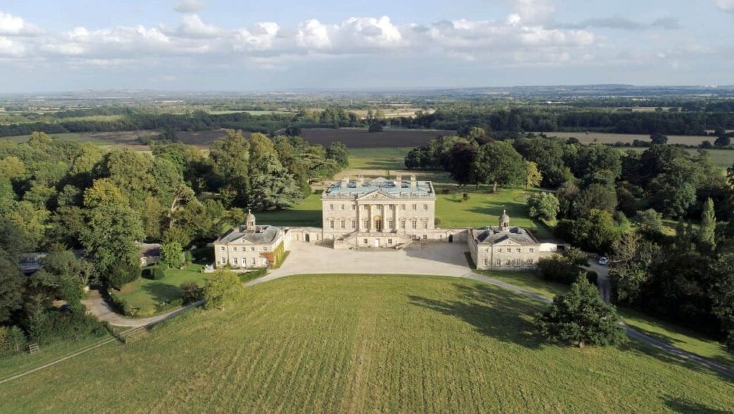 Private: Kirtlington Park, Oxfordshire – Aerial view of grand country house with portico, flanked by pavilions, surrounded by parkland and countryside.