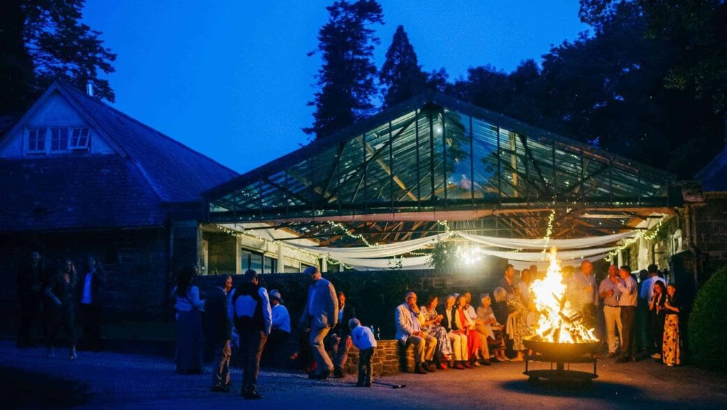 Plas Dinam, Powys – Evening garden party under glass pavilion with central firepit and guests gathered around flames.