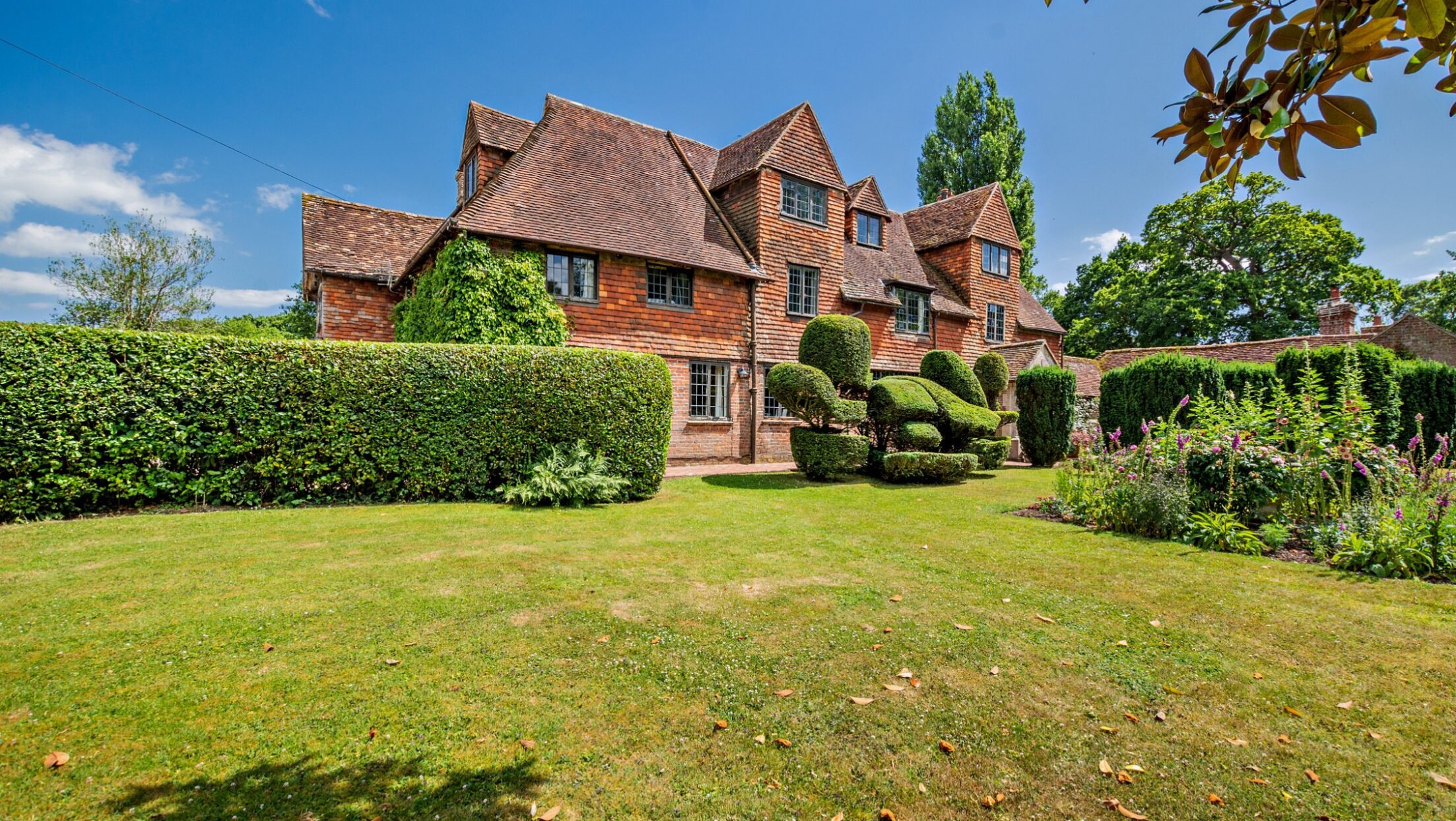 Pekes Manor Estate, East Sussex – Red-brick manor house with gabled roof and topiary-studded lawn bordered by hedges.