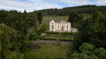 Assynt House, Ross-shire – Cream-rendered castle with towers set in wooded valley with manicured gardens and conifer forest beyond.