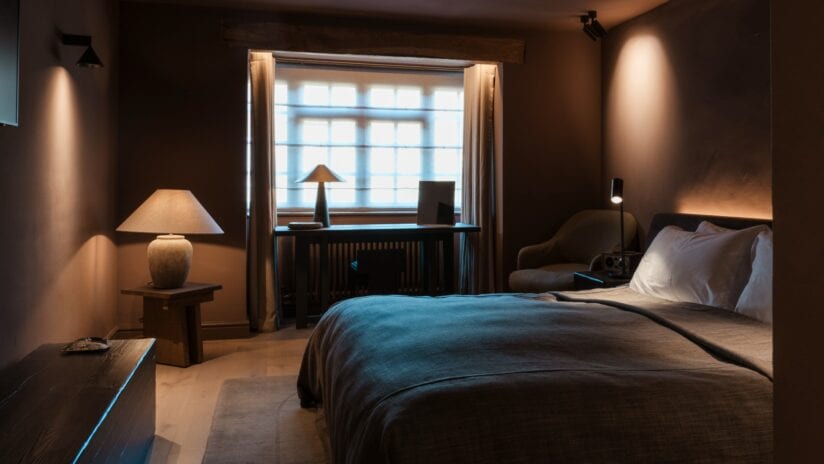 Hyll Hotel, Gloucestershire – Contemporary bedroom with blue upholstered bed, desk by sash window, and warm ambient lighting.