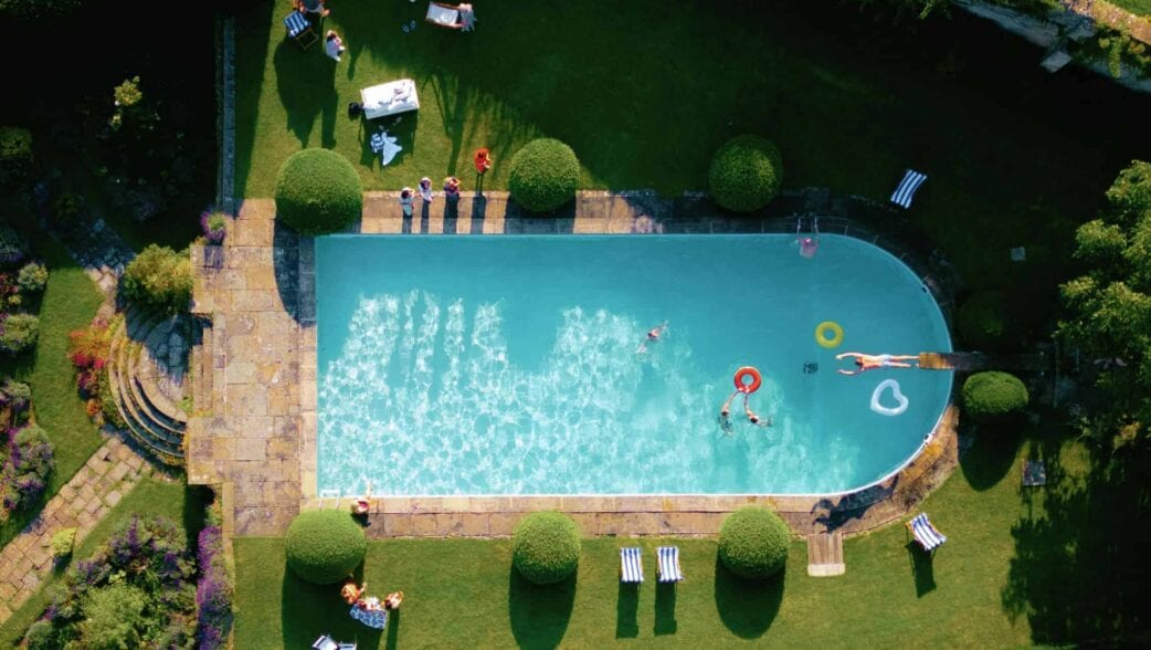 Private: Cornwell Manor, Oxfordshire – Aerial view of heated swimming pool with surrounding terrace, manicured lawns and topiary.