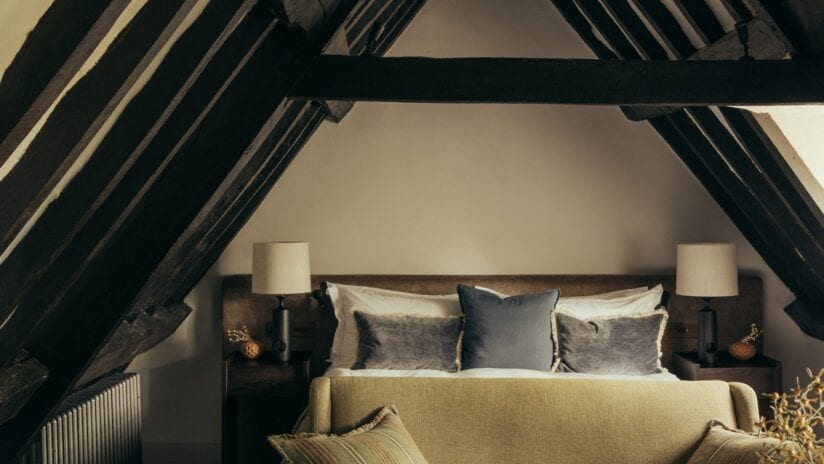 Hyll Hotel, Gloucestershire – Attic bedroom with exposed dark timber beams and cream upholstered bed.