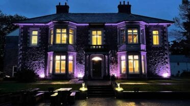 Plas Y Bryn – Stone country house exterior at dusk, illuminated by purple uplighting, with ivy-covered walls and sash windows.