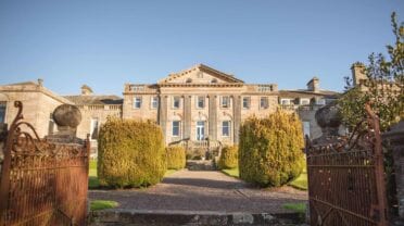 Springkell – Georgian country house with columned façade, gravel drive, and topiary hedges framed by iron gates