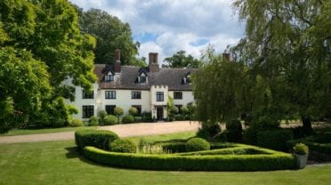 Hill House Norfolk, Norfolk – Cream-rendered country house with black gable windows, brick chimneys, gravel drive, and manicured grounds with topiary hedges.